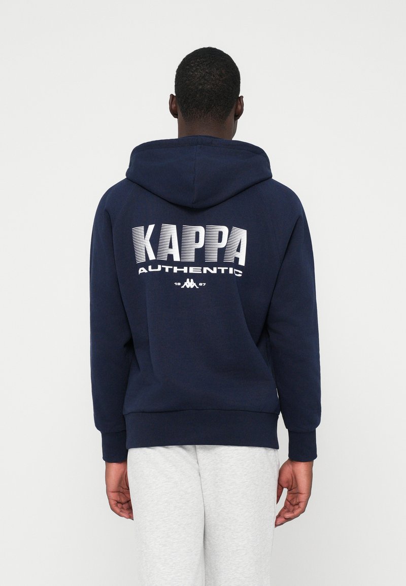 Muž otočený chrbtom, oblečený v tmavomodrej mikine s kapucňou s textom a logom "Kappa Authentic 1967" na chrbte, v kombinácii so svetlosivými teplákmi.