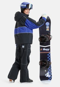 Snowboardzista w czarno-niebieskiej kurtce i czarnych spodniach trzyma deskę snowboardową Roxy. Wyróżniają się niebieskie akcenty oraz gogle ochronne.