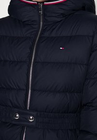 Doudoune bleu marine avec coutures horizontales, fermeture éclair, taille cintrée avec boutons-pression et capuche doublée avec des accents rouges et blancs.