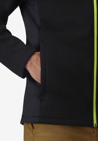 Mano nella tasca di una giacca nera testurizzata con zip verde neon, abbinata a pantaloni marroni su uno sfondo semplice.