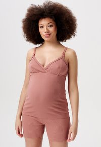 Kobieta w ciąży z kręconymi włosami, ubrana w top bez rękawów w kolorze przybrudzonego różu i dopasowane szorty, stojąca na jednolitym tle.