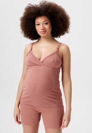 Zwangere vrouw met krullend haar, gekleed in een stofroze ribgebreide mouwloze top en bijpassende shorts, staande tegen een effen achtergrond.