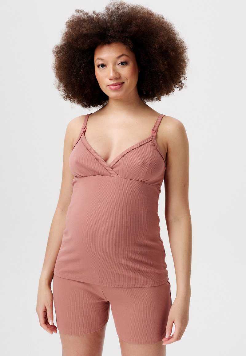 Kobieta w ciąży z kręconymi włosami, ubrana w top bez rękawów w kolorze przybrudzonego różu i dopasowane szorty, stojąca na jednolitym tle.