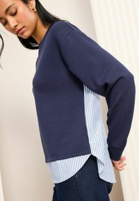 Felpa navy con maniche lunghe e un pannello laterale a righe blu e bianche. Presenta un colletto tondo e una vestibilità rilassata, con un look a strati.
