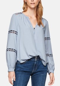 Vaaleansininen paita, jossa on V-pääntie, pitkät hihat ja koristeellinen musta kirjonta hihoissa. Pehmeä kangas, jossa on hieman kiiltoa.