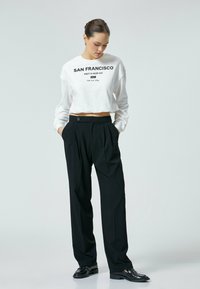 Witte cropped sweatshirt met "San Francisco" opdruk, gecombineerd met zwarte high-waisted, geplisseerde broeken en glanzende zwarte loafers.