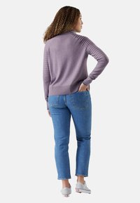 Donna con i capelli ricci, che indossa un maglione a coste di colore lilla, jeans blu e sneakers bianche, in piedi con una mano in tasca, rivolta di spalle.