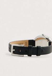 Μαύρη δερμάτινη λουρίδα ρολογιού με ασημένια μεταλλική πόρπη, που διαθέτει στρογγυλό ασημένιο καντράν ρολογιού και διακριτική υφή στη λουρίδα.