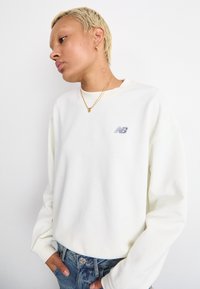 Sudadera color crema con puños de canalé, que presenta un pequeño logo gris "NB" en el pecho. Llevada con jeans de mezclilla azul.