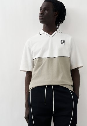 Homme à la peau foncée et aux dreadlocks portant un polo blanc et beige à manches courtes et un pantalon noir avec des cordons blancs et des rayures.