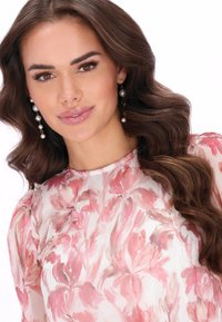 Kwiatowa bluzka z delikatnego, półprzezroczystego materiału w jasnoróżowym kolorze z ciemniejszymi różowymi kwiatami. Modelka ma na sobie perłowe kolczyki wiszące na tle falowanych brązowych włosów.