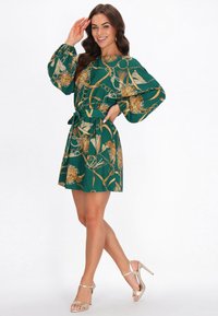 Zöld mintás ruha arany virág- és láncmintával, hosszú, puffos ujjakkal, derékrésznél szűkített, és bővülő szoknyával. Fémes magassarkúval viselve.