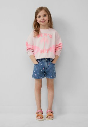 Sweatshirt rose clair avec le graphique "SUN'S OUT", associé à un short en denim bleu orné de motifs de fruits colorés et des sandales roses.