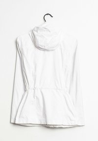 Vit lättviktsjacka med huva, utrustad med dragsko i midjan, elastiska ärmslut och en ren, jämn textur. Hänger på en svart krok.