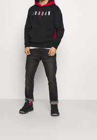Felpa nera con accenti rossi, con la scritta "JORDAN" in lettere bianche. Jeans scuri e sneaker nere con dettagli rossi. Vestibilità e design casual.