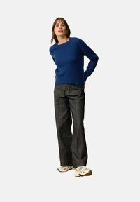 Niebieski sweter dzianinowy z okrągłym dekoltem, połączony z ciemnymi, luźnymi dżinsami i beżowymi trampkami. Tło jest białe.