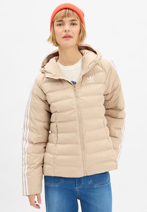 Giovane donna che indossa un piumino beige Adidas con strisce bianche sulle maniche, jeans blu e un berretto di lana arancione, in piedi su uno sfondo bianco.