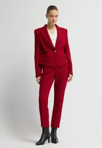 Roter maßgeschneiderter Anzug mit strukturierten Schultern, Ein-Knopf-Verschluss und Fronttaschen. Kombiniert mit einer weißen Bluse und schwarzen Knöchelstiefeln.