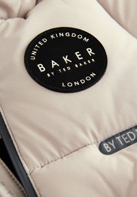 Beige puffer-jakki, jonka kankaassa on musta pyöreä etiketti, jossa lukee "BAKER BY TED BAKER LONDON". Suojaava pinta, hienovarainen kiilto ja kevyt suunnittelu.