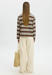Camicia a righe marroni e bianche a maniche lunghe con vestibilità rilassata, abbinata a pantaloni a gamba larga color crema; tiene una borsa tote intrecciata beige.