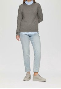 Kvinna i ljusblå randig skjorta, grå tröja, ljusblå jeans och grå plattformssneakers mot en enfärgad bakgrund.