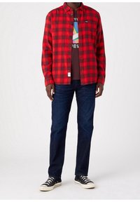 Chemise en flanelle à carreaux rouge et noir, t-shirt graphique marron en dessous, jeans en denim foncé et baskets noires avec semelles blanches.