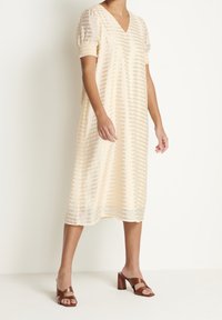 Cremfarbendes Kleid mit einem V-Ausschnitt, kurzen Puffärmeln und vertikaler Streifenstruktur. Getragen mit braunen Blockabsatzsandalen.