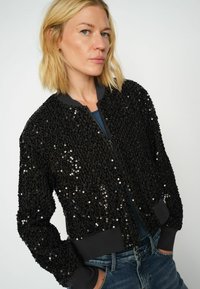 Crna bomber jakna s šljokicama, sa zatvaračem sprijeda, rebrastim manžetama i pojasom. Tkanina je teksturirana s sjajnim završetkom.
