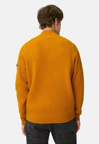 Rippstrickpullover in senfgelb, mit hohem Kragen und langen Ärmeln, einem dezenten Logo-Detail auf der Rückseite und kontrastierenden dunklen Hosen.