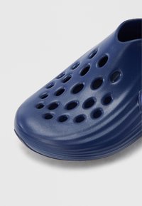 Blauwe synthetische klomp met een gladde textuur, grote ventilatiegaten en een afgerond neusontwerp. Platte zool met gebogen randen.