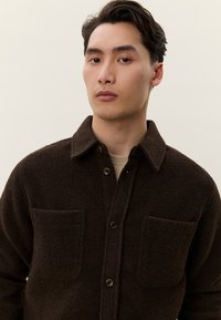 Jaunas vyras su trumpais tamsiais plaukais, dėvintis tamsiai rudos spalvos vilnonę marškinių striukę su dviem krūtinės kišenėmis, stovintis prieš paprastą foną.