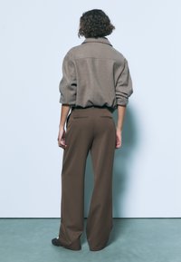 Ruskea ylisuurikokoinen villapaita leveällä kauluksella, yhdistettynä ruskeisiin korkeavyötäröisiin, leveälahkeisiin housuihin, nähtynä takaa.