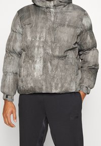 Puffig grå jacka med en texturerad yta, som har huva, blixtlås och elastiska manschetter, tillsammans med svarta byxor.