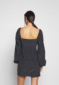 Frau mit dunklem Pferdeschwanz, die ein schwarzes Kleid mit weißen Punkten, quadratischem offenen Rücken und langen puffärmeligen Ärmeln trägt, mit dem Rücken zur Kamera.