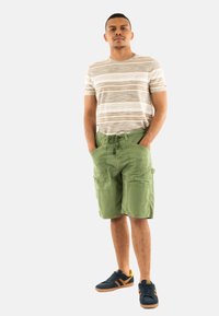 T-shirt rayé beige et marron, shorts cargo verts avec poches, et baskets foncées avec accents orange, se tenant devant un fond blanc.