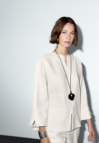 Chemise en lin beige clair avec fermeture à boutons et fentes latérales, associée à un bracelet en argent texturé et un pendentif en forme de cœur noir sur un cordon.