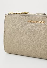 Beige läderplånbok med en texturerad yta, blixtlås och detalj med guldtonat "MICHAEL KORS"-logotyp på framsidan. Kompakt och rektangulär design.