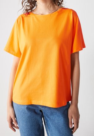 Person iført en ensfarvet klar orange kortærmet skjorte og blå jeans foran en ensfarvet lys baggrund.