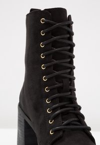 Bottine en daim noir à bout arrondi, talon bloc épais, œillets dorés et lacets noirs. Présente une surface texturée et un design ajusté.