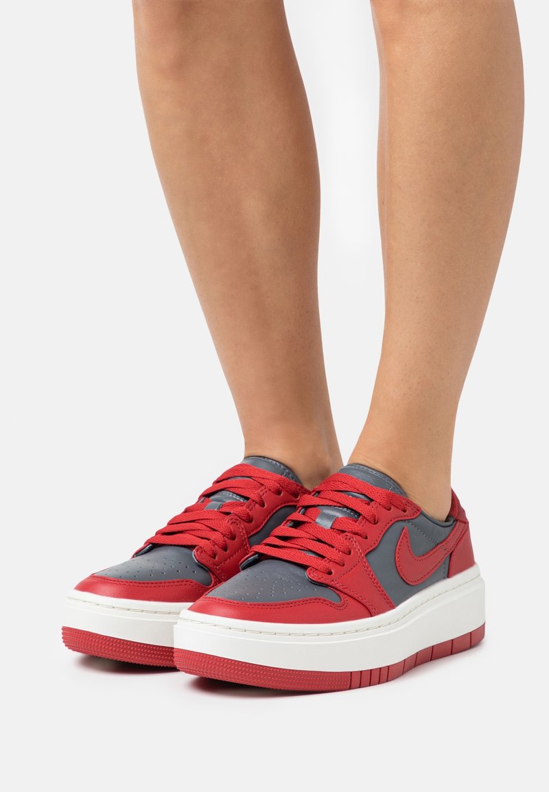Jambes portant des baskets basses Nike rouges et grises avec des semelles blanches épaisses sur fond blanc.