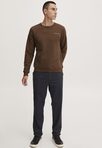 Brun sweatshirt med lange ermer og rund hals, med en liten hvit logo, kombinert med mørke, teksturerte bukser og svarte sneakers.