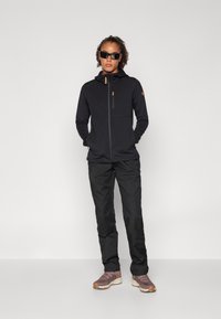 Giacca nera con zip e cappuccio, con una tasca frontale e dettagli color beige. Abbinata a pantaloni neri e scarpe grigio-marroni. Il modello indossa occhiali da sole.