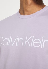Camiseta de algodón lavanda con cuello redondo, que presenta un prominente logo blanco de "Calvin Klein" impreso en el pecho.