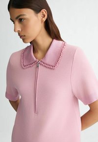 Růžový pletený polo tričko s uzávěrem na zip, texturovanou tkaninou a korálkovým designem límce. Krátké rukávy dotvářejí vzhled.