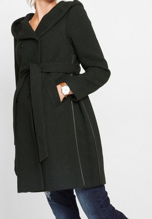 Nainen, joka pitää tummaa kalanruotokuvioista, vyöllä varustettua takkia, jossa on huppu, käsi taskussa, yhdistettynä sinisiin farkkuihin ja ruskean nahkahihnan rannekelloon.