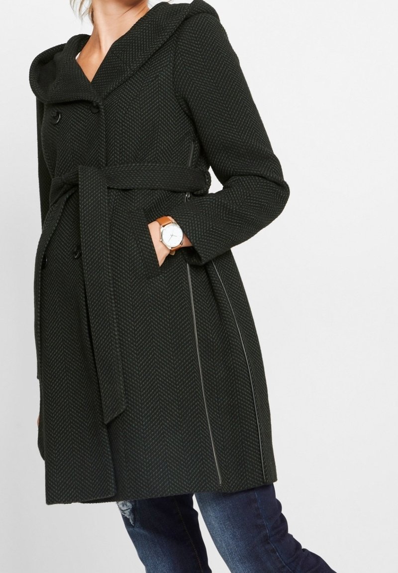 Femme portant un manteau sombre à chevrons ceinturé avec capuche, main dans la poche, associé à un jean bleu et une montre bracelet en cuir marron.