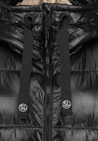 Veste matelassée noire devant avec une fermeture éclair centrale et deux larges cordons noirs ornés de logos blancs "SL Studio" près des extrémités.