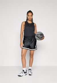 Kvinna i svart ärmlös topp, svarta basketshorts, vita sneakers, som håller en svart och grå basket mot en enkel bakgrund.