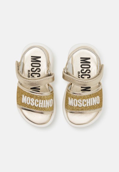 Zelta sandales ar divām siksnām: viena spīdīga zelta, otra ar zelta mirdzumu, uz kuras ir vārds "MOSCHINO" baltā krāsā. Mīksts balts zole.