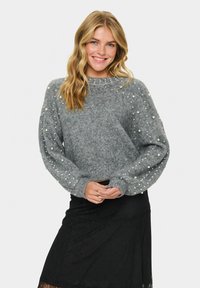 Szürke kötött pulóver gyöngy díszítéssel az ujjakon és bordázott aljjal. Kerek nyakkivágású, és enyhén cropped fazonú. Fekete szoknyával párosítva.